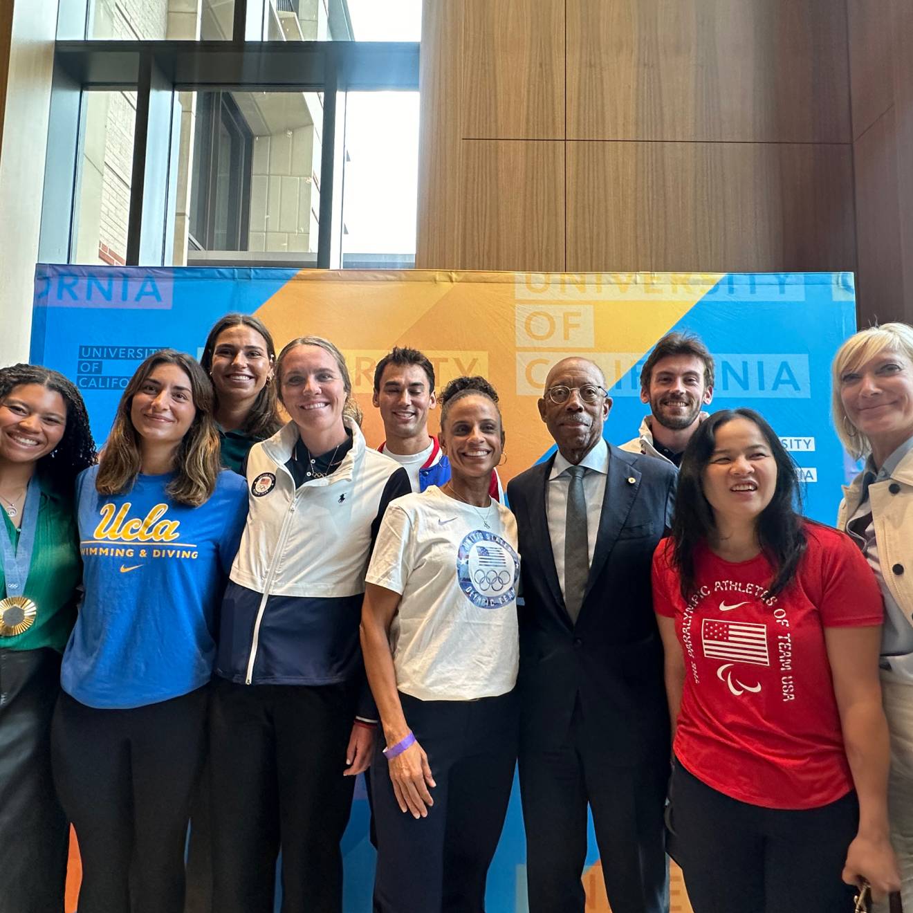 Olympian and Paralympian athletes stand with President Drake and Board of Regents Chair Janet Reilly in front of a University of California backdrop for photos