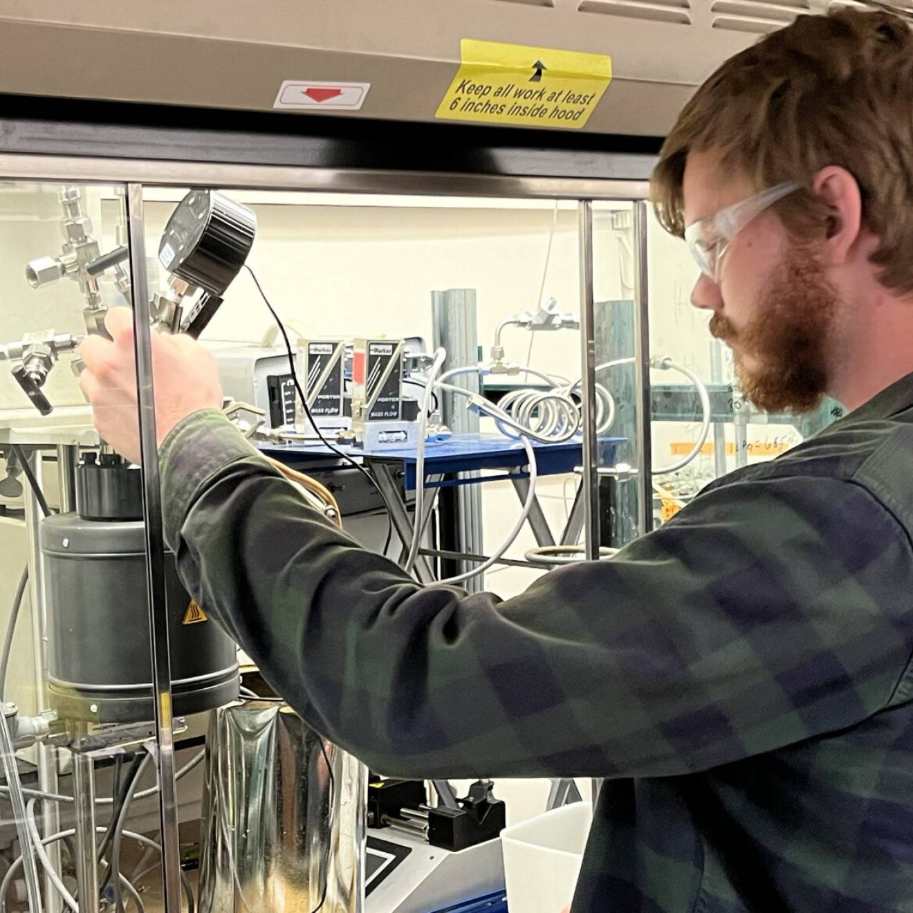 A young bearded man with red hair and protective glasses operates a small machine