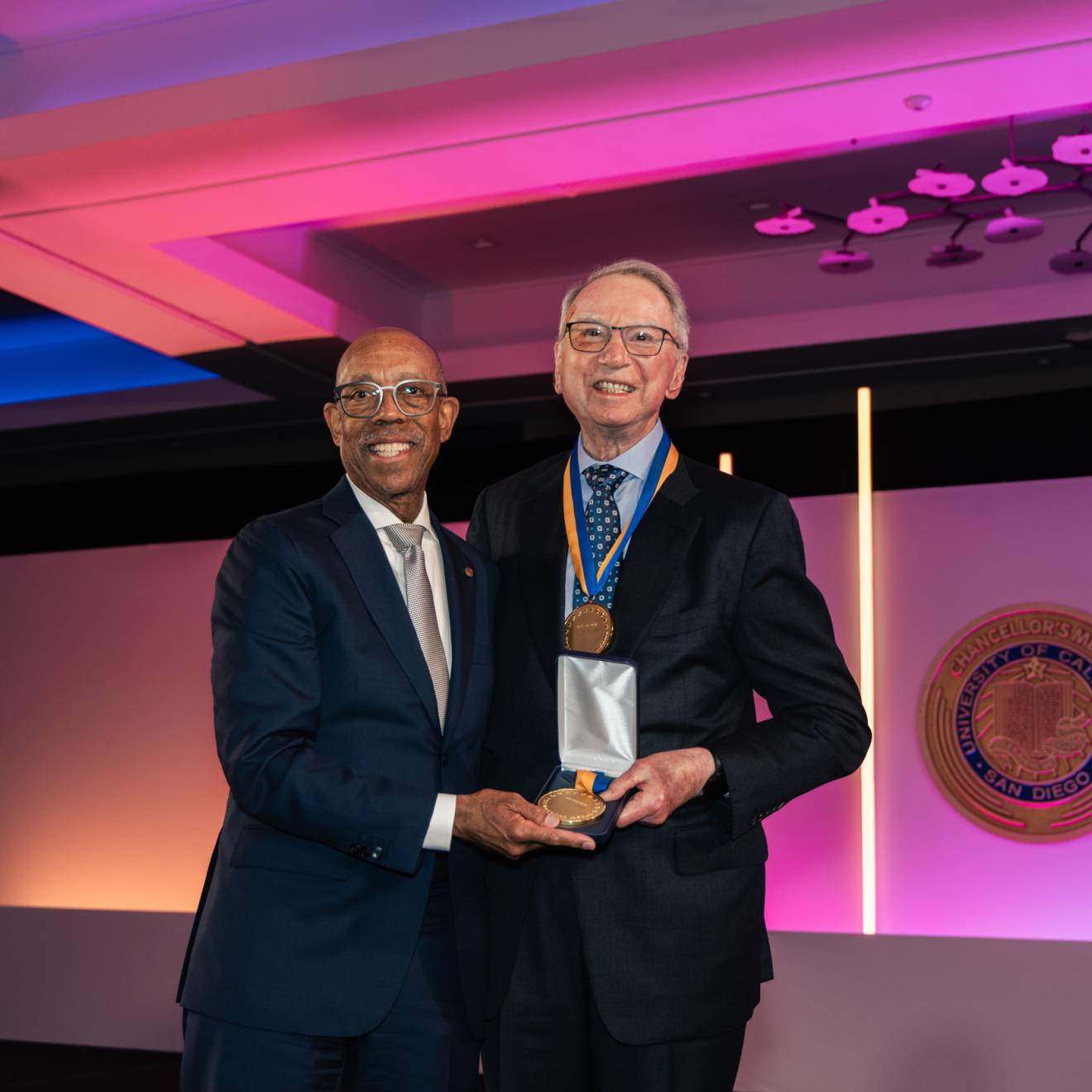 President Drake and Irwin Jacobs pose together in a room lit with purple lighting while Jacobs wears a medal and holds another