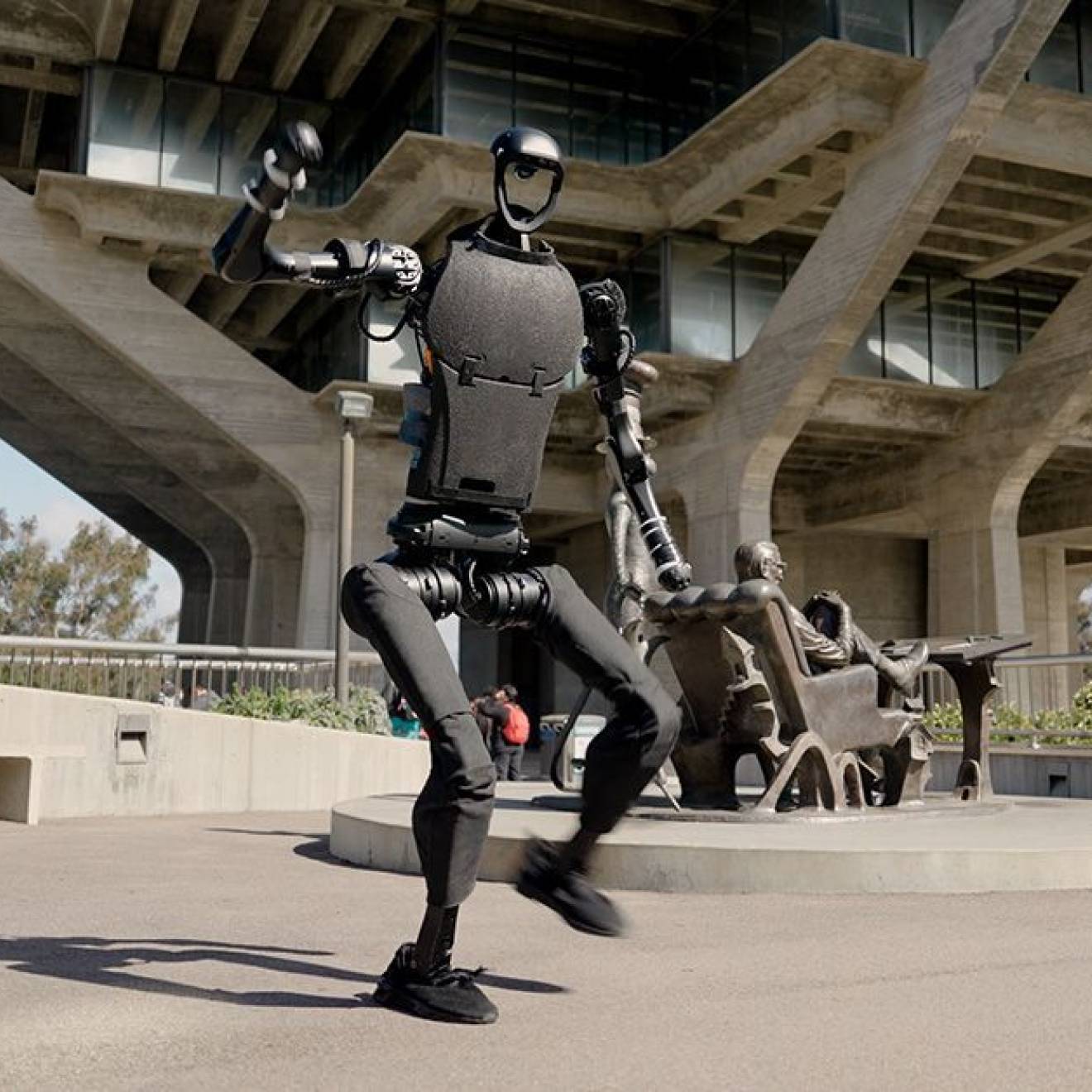 A humanoid robot on the UC San Diego campus in mid-dance move, Geisel Library visible behind