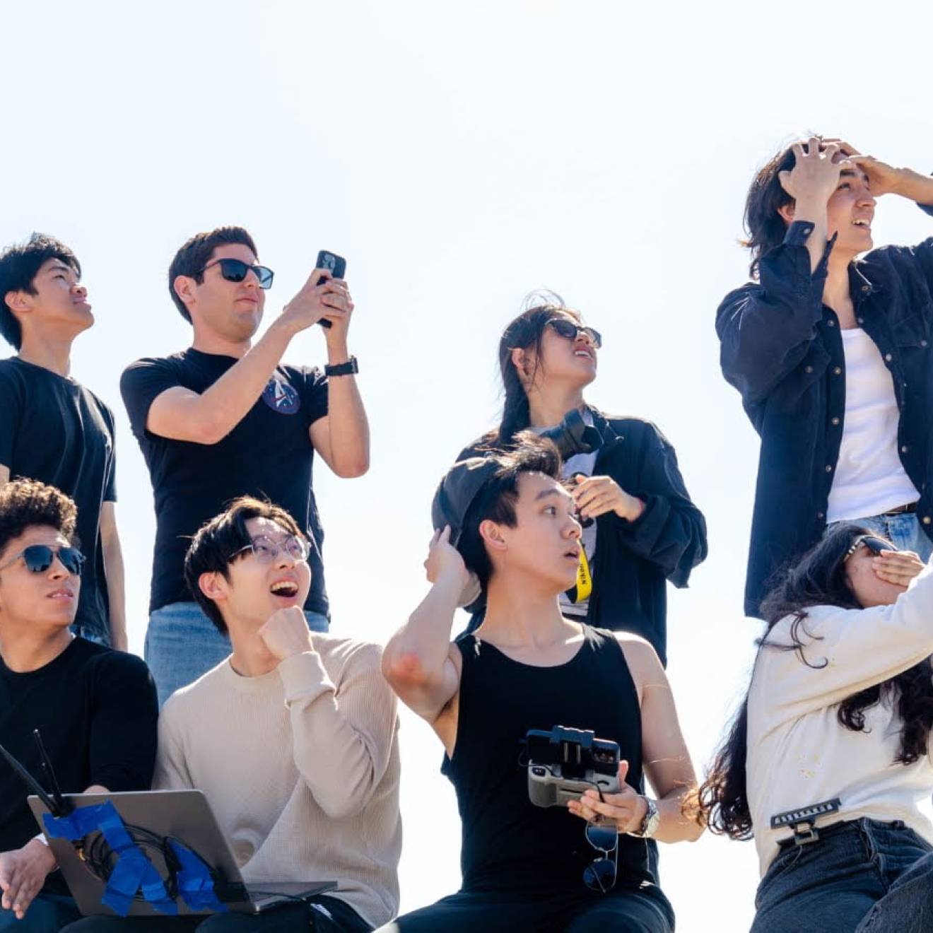 A group of eight people looking at something off camera, four standing, four seated, some gasping, a few with phones recording something, some looking shocked (presumably watching a rocket)