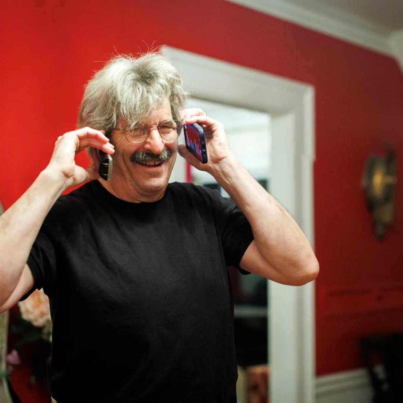 A happy man with white hair and a salt and pepper mustache answers two phones at the same time, one on each ear, laughing