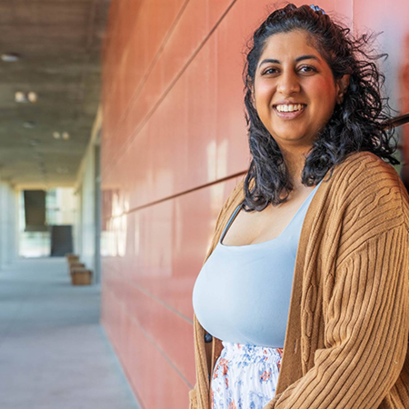 Woman with shoulder-length dark curly hair smiles while standing in an outdoor UC Merced campus corridor