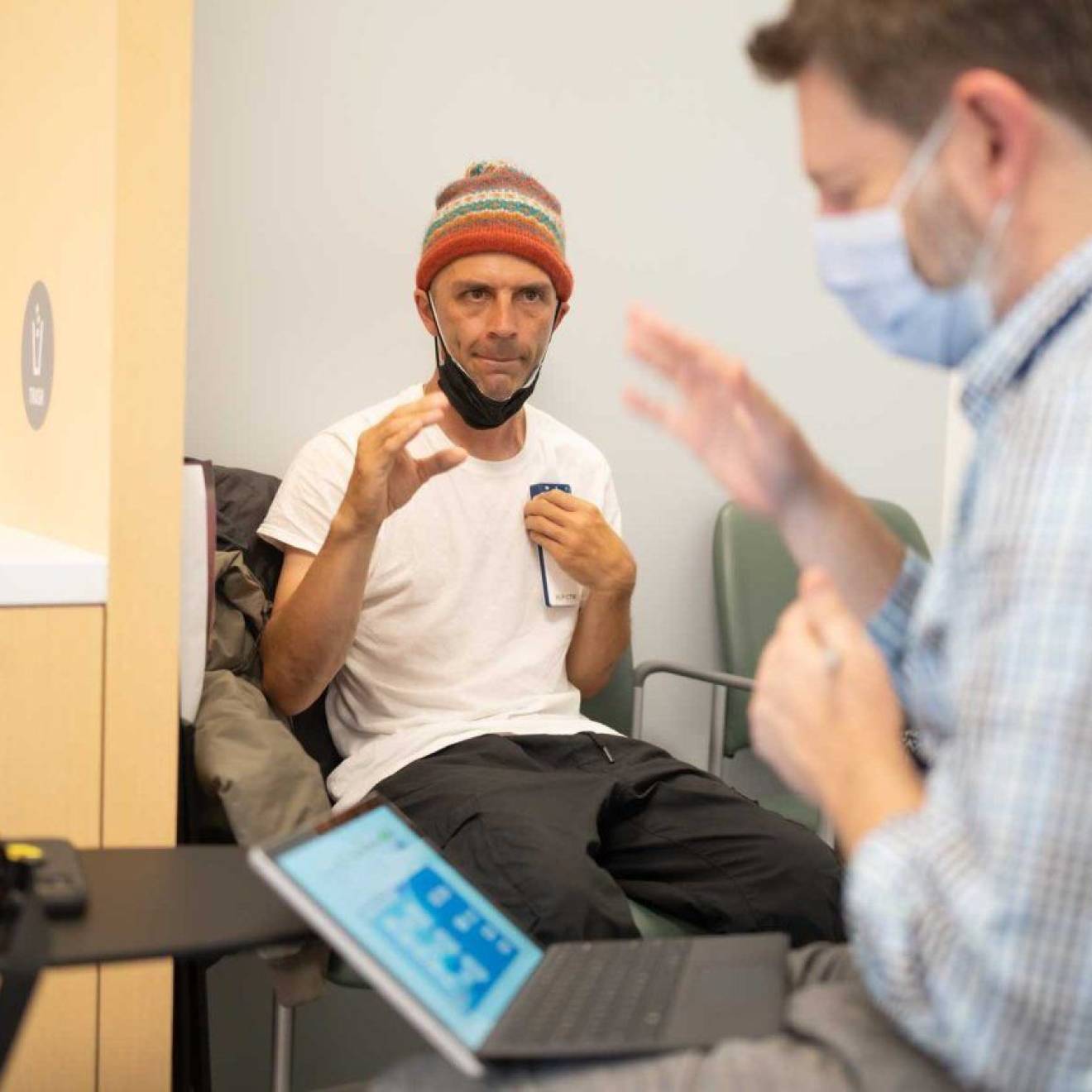 New Parkinson’s therapy In the background, in focus, a man in a beanie makes a gesture with his hand as if to grasp. In the foreground, a doctor in a mask in front of a computer screen makes the same gesture