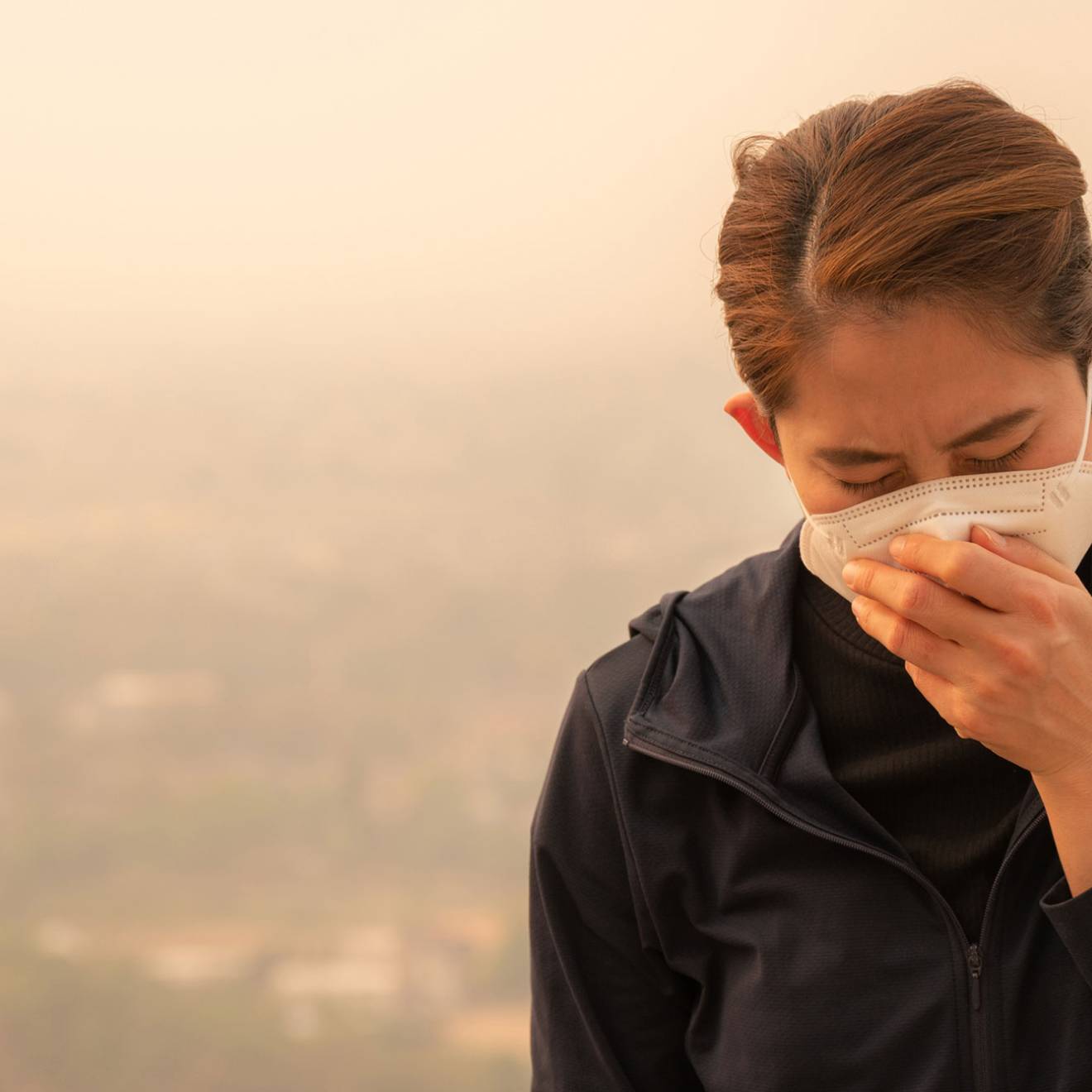 Woman wearing a face mask and coughing into her hand. They sky behind her is hazy with smoke.