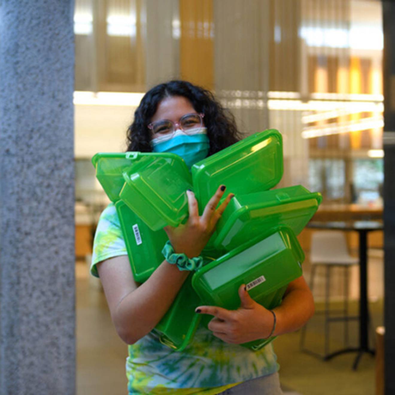 Woman in a mask carrying an armload of green reuseable food containers