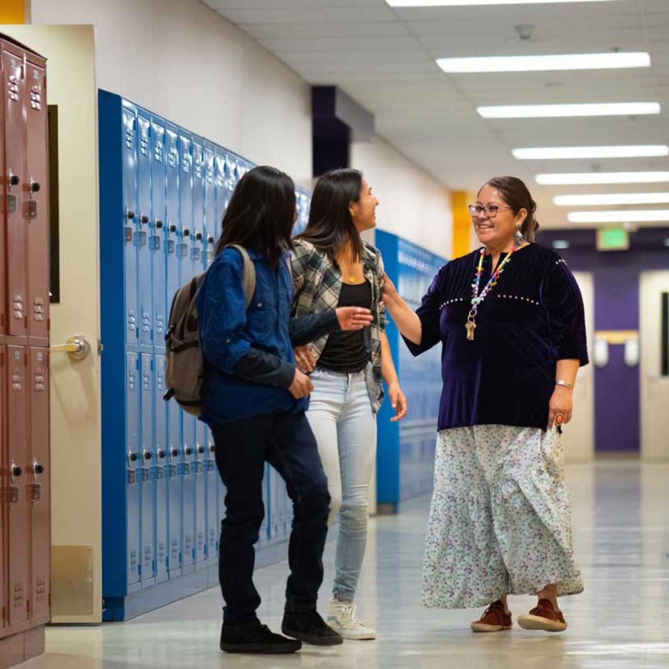 An adult talking with two high school students in a school hallway, with rows of lockers in the background