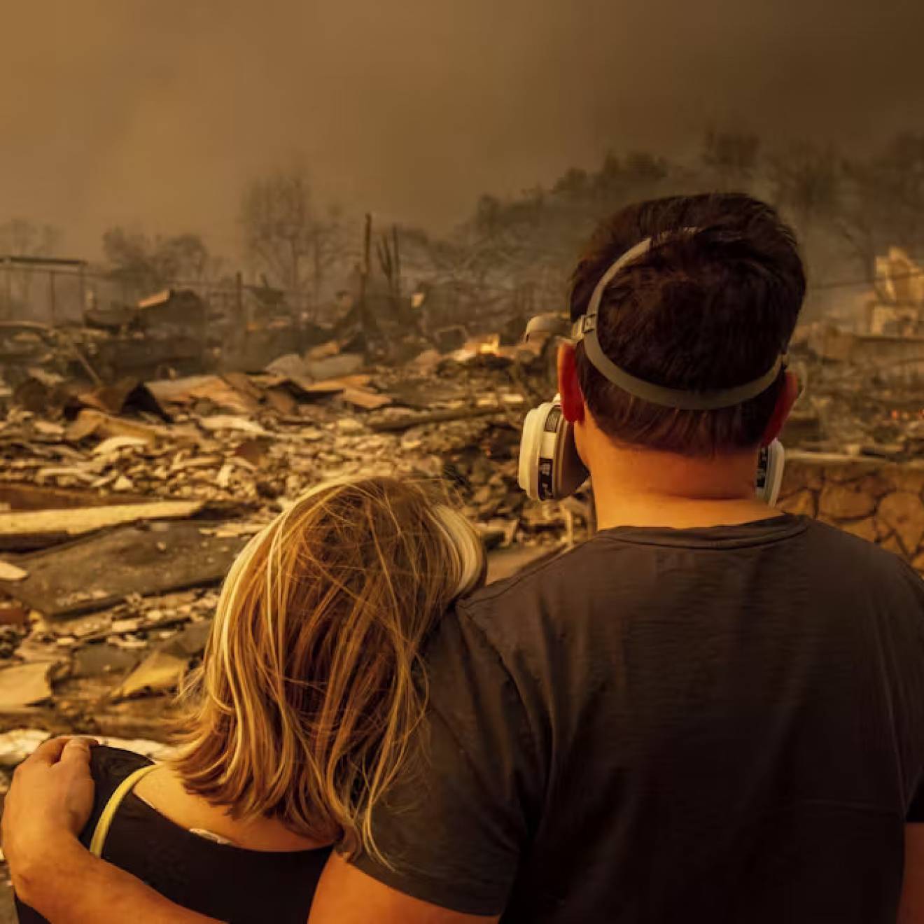 A young man in a mask and woman, photographed from behind, look on at an obliterated house with smoke all around