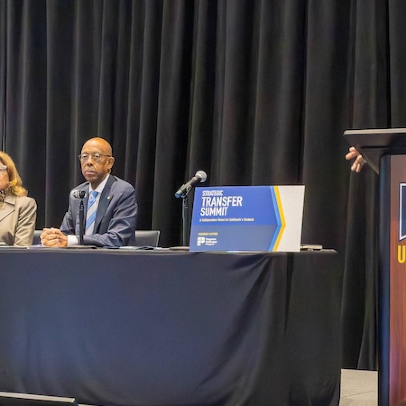 At a table, three people sit in front of microphones, from left: California Community Colleges Chancellor Sonya Christian, California State University Chancellor Mildred Garcia, and UC President Michael Drake; at a lectern, UC Merced Chancellor Juan Sánchez Muñoz stands. Audience is visible a bit in the front of the photo 