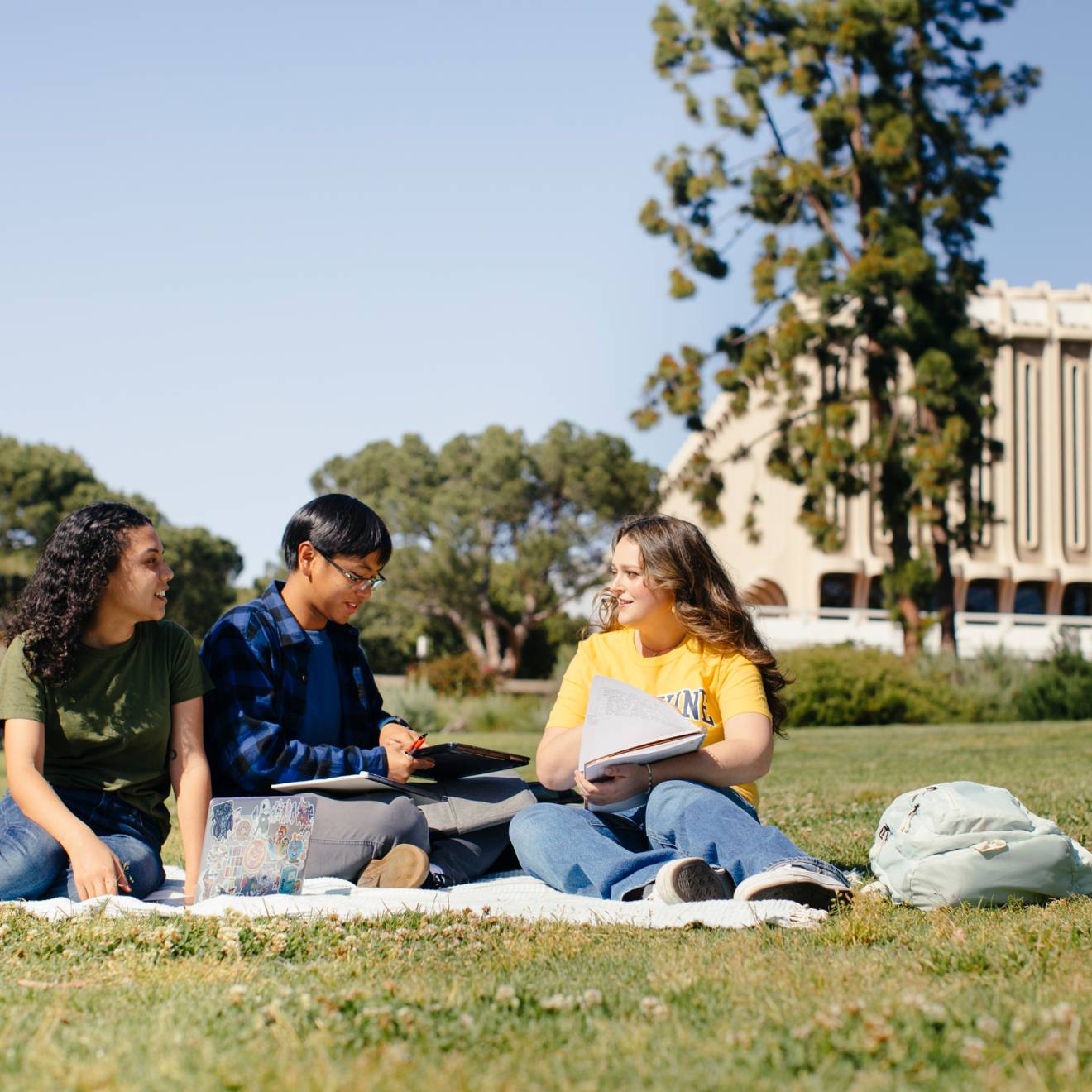 Three students study together on a blanket on the lawn of the UC Irvine campus, tall trees and buildings visible in the background