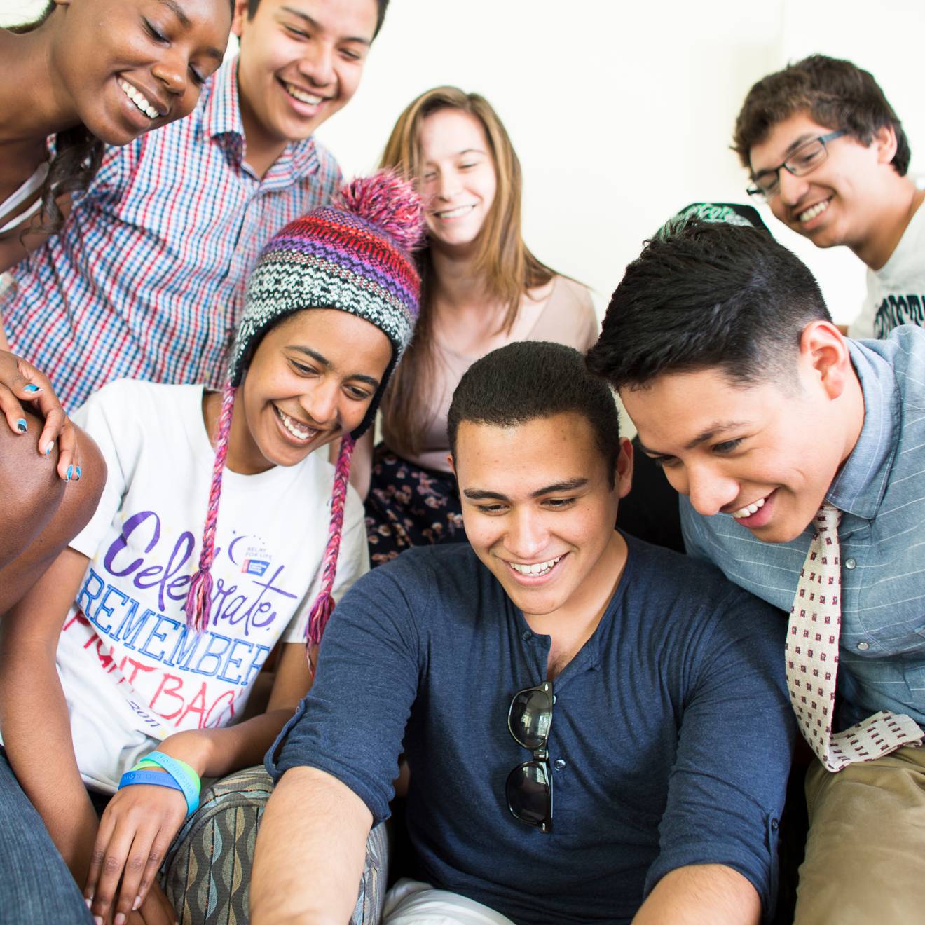 A diverse group of students gather together to look down at something out of frame, like a computer