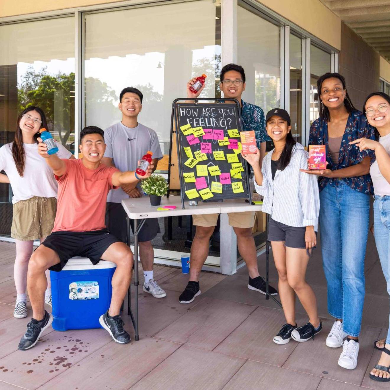 A group of smiling students in front of a dorm with bottles of water and a board that says How Are You Feeling and a number of responses