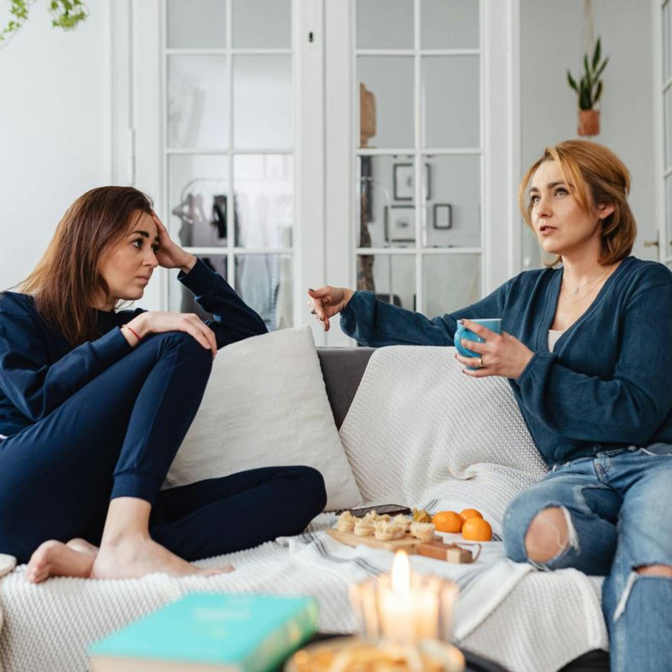 Two women sit on a couch drinking tea with a snack tray between them. The woman on the right is speaking and the woman on the left is listening.