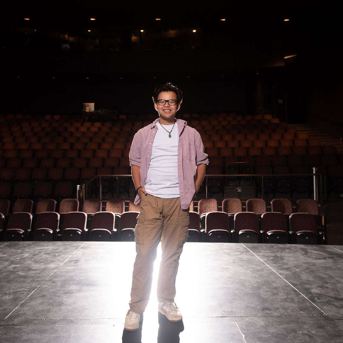 A smiling man standing on an empty theater stage under dramatic lighting