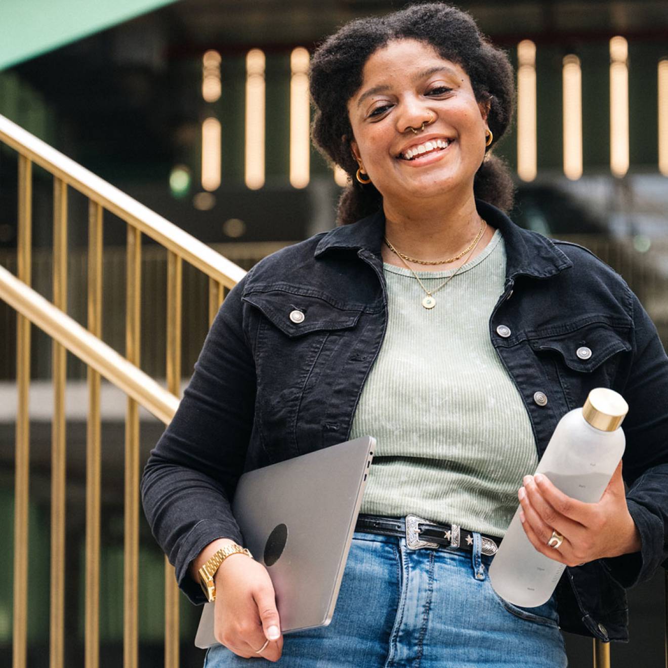 A college student smiles for the camera on a staircase in a university building, holding a reusable water bottle and a laptop