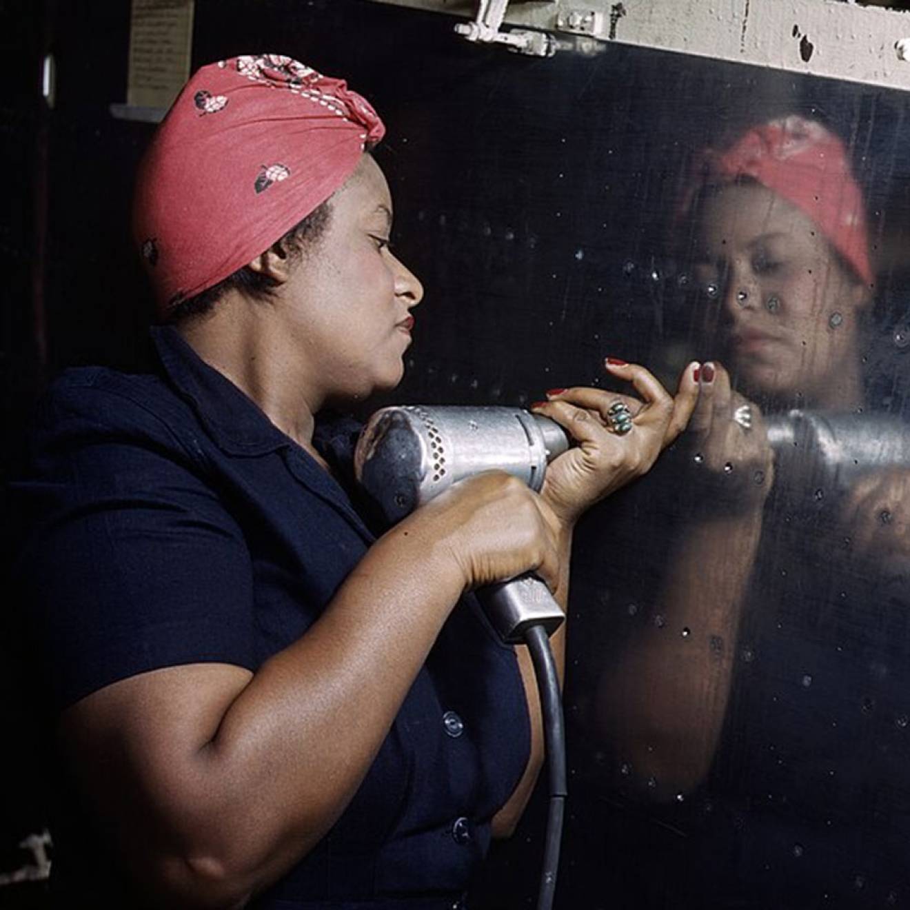 A Black woman uses a rivet gun on a reflective steel panel, wearing a red headscarf and a blue jumpsuit,