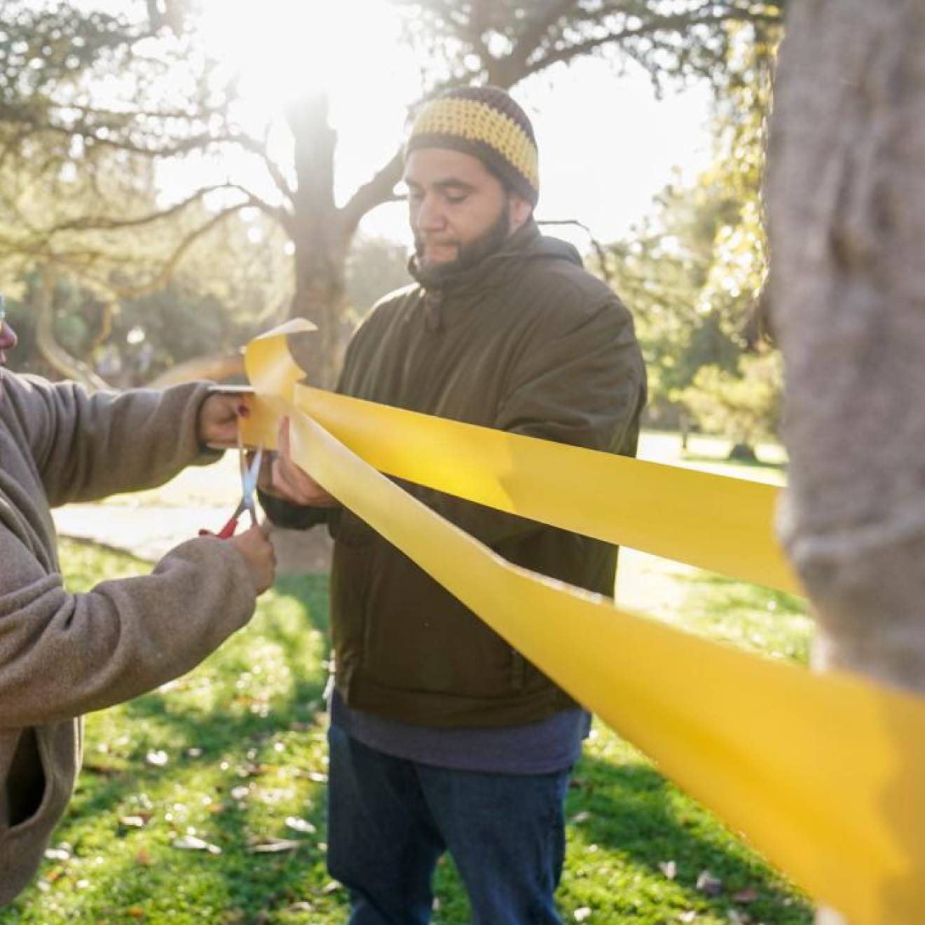 A woman cuts a long yellow ribbon tied to a tree a man is holding